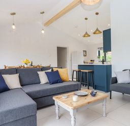 An open living space with gray sofas, a wooden coffee table, and a blue kitchen area at Woodpecker Lodge in Butterleigh