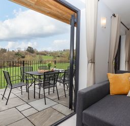 A patio dining table with six chairs and flowers overlooking green fields next to a room with a gray sofa and curtains at Woodpecker Lodge in Butterleigh