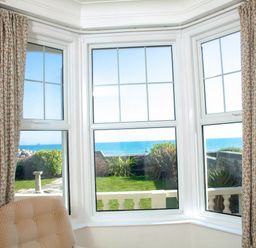 A window with three panes overlooking a garden and the sea with patterned curtains and a beige chair at Weymouth Bay Apartment A in Weymouth