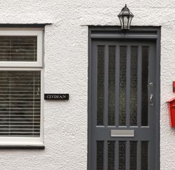 A grey front door with glass panels next to a window with blinds a red mailbox and a black house nameplate reading Clydfan on a white textured wall at Clydfan in Caernarfon