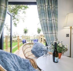 A small table with wine bottle and two wine glasses between two chairs with blue blankets next to a lamp and open door leading to an outdoor deck with chairs at High Rigg Shepherd's Sunset in Brampton Cumbria