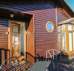 An outdoor patio with a glass table and metal chairs outside a wooden cabin with a door and circular window at Badgers Retreat in Tunstall near Hipswell