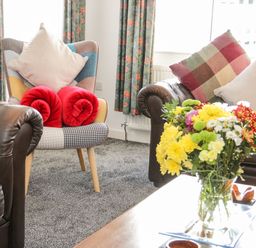 A living room with leather sofas and a patterned armchair with cushions and red rolled blankets a vase of flowers on a coffee table in Gwersyllt Benllech