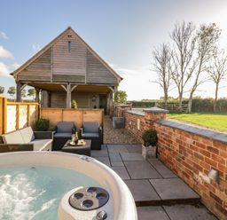 An outdoor patio with a hot tub seating area and a wooden building with a grassy field beyond at The Hyde in Armscote near Newbold on Stour
