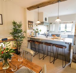 A kitchen with a breakfast bar and stools next to a dining table with flowers and place settings at The Hyde in Armscote near Newbold on Stour