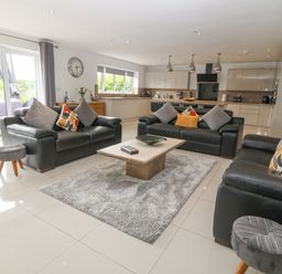 A living room with three black leather sofas and a coffee table on a rug with a kitchen in the background at Berllan in Llanbedrgoch near Benllech