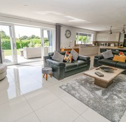 An open living room and kitchen area with black sofas a round chair a coffee table on a rug and large sliding glass doors at Berllan in Llanbedrgoch near Benllech