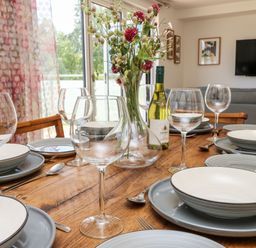 A dining table set with plates, wine glasses, a vase with flowers, and a bottle of wine in a room with a television and curtains at Cava in Seaview