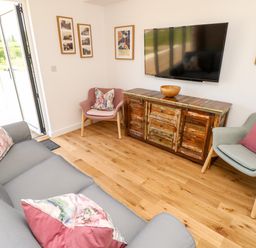 A living room with a gray sofa and pink and floral cushions a wooden cabinet with a bowl and a wall-mounted TV two chairs with cushions and framed pictures on the wall at Chianti in Seaview