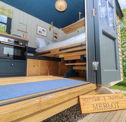 A small interior space with a bed, blue cabinets, an oven, wooden storage, and an open door showing greenery outside at Merlot in Pondwell near Seaview