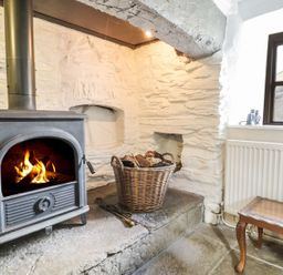 A wood burning stove with fire in a stone room next to a basket of firewood and a wooden table at Tan Y Garth Cottage in Glyndyfrdwy near Llangollen