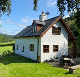 A white cottage with a slate roof and wooden windows in a green grassy area with outdoor seating at Tan Y Garth Cottage in Glyndyfrdwy near Llangollen