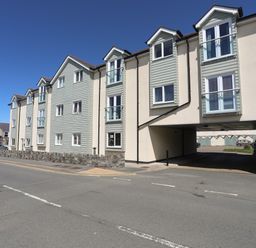 A street view of a multi-story residential building with light green and cream facades and an archway entrance at 8 Pen Llanw Tides Reach in Rhosneigr