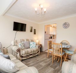 A living room with sofas a wall-mounted television a dining table with chairs and a kitchen in the background at Chalet 12 in Hemsby
