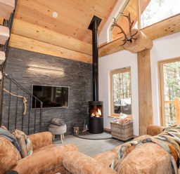 A living room with a wood burning stove a flat screen TV and a staircase with wooden beams and large windows at Pine Marten Lodge Dulnain Bridge near Nethy Bridge