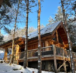 A wooden cabin in a snowy forest with a deck and railing at Pine Marten Lodge in Dulnain Bridge near Nethy Bridge