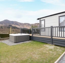 An outdoor area with a hot tub next to a deck attached to a white building with mountains in the background at Red Kite Lodge in St Fillans