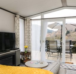 A living room with a television a round coffee table and sliding glass doors opening to a balcony with chairs and an umbrella at Red Kite Lodge in St Fillans