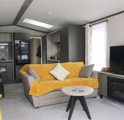 A living room with yellow covers on sofas a round wooden coffee table and a TV with fireplace at Red Kite Lodge in St Fillans