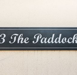 A black rectangular house sign with white text reading 3 The Paddock on a textured beige wall at 3 The Paddock in Benthall near Beadnell