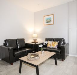 A living room with two black sofas a wooden coffee table with a wicker tray and flowers a lamp and wall art at Llety Pen Deitsh 1 in Caernarfon