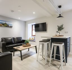 A living room with black leather sofas a wooden coffee table a wall-mounted TV a kitchen island with three bar stools and two windows at Llety Pen Deitsh 2 in Caernarfon