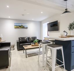 A living room with two black leather sofas a wooden coffee table a kitchen island with stools and a wall-mounted TV at Llety Pen Deitsh 2 in Caernarfon