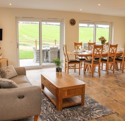 A living area with a sofa, coffee table, dining table with chairs, and sliding glass doors overlooking a green field at Holly Lodge in Forden near Montgomery