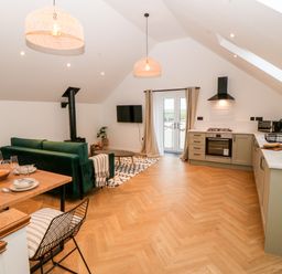 An open plan kitchen and living area with a dining table a green sofa a wall-mounted TV and kitchen cabinets at Sgubor in Llangefni