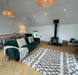 A living room with a green sofa and patterned rug near a wood stove and a dining table at Sgubor in Llangefni