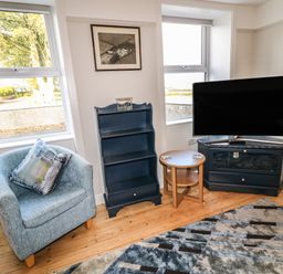 A corner of a living room with a blue armchair, a small wooden round table, a blue shelving unit, a flat screen TV on a blue TV stand, and two windows at Grannys in Bushmills County Antrim