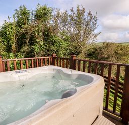 An outdoor hot tub on a wooden deck with wooden railing surrounded by trees and fields at Orchard View in Bossiney near Tintagel