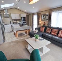 An open living room and kitchen area with a dark sectional sofa with patterned pillows a light wooden coffee table with a plant a dining table with a tablecloth and wooden benches and kitchen cabinets and appliances at Windermere View in Pony Meadow 37 White Cross Bay near Windermere