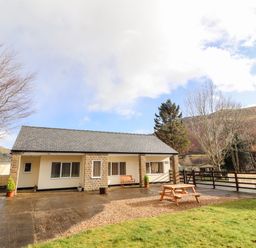 A single story building with outdoor bench and picnic table on a paved and gravel area with grass and trees surrounding at Awel Y Berwyn in Llangynog