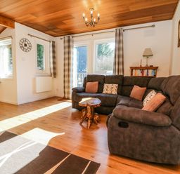 A living room with a large sectional sofa, small wooden table, bookshelves, and windows with plaid curtains at Stonewood Lodge in Woolsery