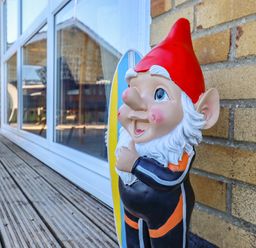 A garden gnome with a red hat and white beard leaning against a brick wall on a wooden deck outside a building with large windows at Bungalow 14 in Corton