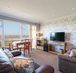 A living room with two sofas a dining table and chairs a television on a wooden stand and large windows overlooking a grassy area at Bungalow 14 in Corton