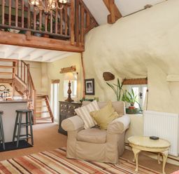 A living room with a beige armchair and pillows a small table and plants near windows and stairs at The Byre in Monkleigh near Great Torrington