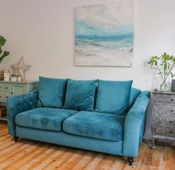 A teal sofa with three cushions between a green sideboard with plants and decorative items and a patterned chest with a vase of flowers in a room at The Lazy Seal in Spittal