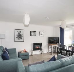 A living room with blue sofas a television a dining table and windows at 5 Ger Y Nant in Llanbedrog