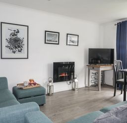 A living room with blue couches a wall-mounted fireplace a television on a corner table and framed pictures on the wall at 5 Ger Y Nant in Llanbedrog