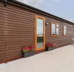 The exterior side of a wooden lodge with a door, two windows, flower boxes, and two lounge chairs on a concrete patio