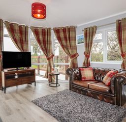 A living room with a leather sofa a television on a wooden stand and plaid curtains at Sycamore Lodge in Porkellis near Helston