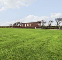 A wooden cabin on a grassy field with a fenced area and trees in the background at Sycamore Lodge in Porkellis near Helston