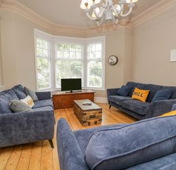 A living room with three blue sofas a wooden floor a wooden chest used as a coffee table a TV on a low wooden cabinet and large bay windows at Hollywood in Windermere
