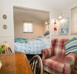 A room with a wooden table and chairs a red plaid armchair a bed with blue checked bedding and windows with blinds at Seashore Shepherds Hut @ Moat Farm in Aldingham near Ulverston
