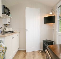 A small kitchen area with cabinets a microwave kettle and toaster a door a wall mounted television a small stove and a table near a window at Seashore Shepherds Hut @ Moat Farm in Aldingham near Ulverston