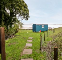 A blue shepherds hut on grass near a fenced area with trees and a view of water at Seashore Shepherds Hut @ Moat Farm in Aldingham near Ulverston