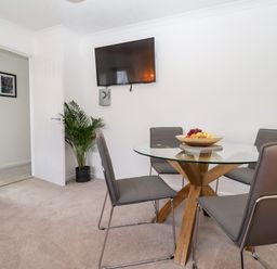 A dining area with a glass table and gray chairs a wall-mounted tv a plant and a wooden shelf at 3 Granbrook Lane in Mickleton Gloucestershire