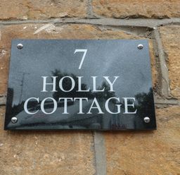 A black house number sign with white text reading 7 Holly Cottage on a stone wall at Holly Cottage in West Coker
